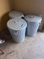 Three galvanized steel trash cans, each with a lid and metal handles, shown from front angle against a wooden wall.