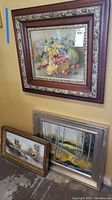 Three framed paintings leaning against a wall. Features a still life fruit painting, a landscape, and a large oil painting with a gold frame.