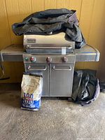 Weber Genesis stainless steel barbecue grill with two control knobs, grill cover draped over it, extra grill cover folded beside, and bag of charcoal briquettes in front