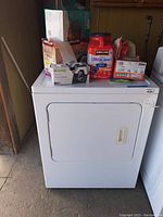 Front view of white Whirlpool electric dryer with assorted laundry supplies on top including detergent, dryer sheets, trash bags, and paper towels