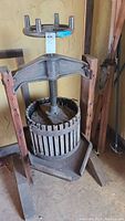Front view of the wine press showing wooden frame, vertical slatted basket, and metal screw with wooden handle.