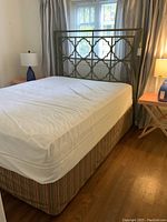 Photo showing entire bed setup with mattress, box spring and gold-tone decorative metal headboard near window with curtains and lamps on bedside tables.