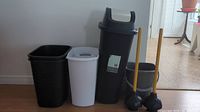 Six waste bins of different heights, one with a lid, two wooden-handled plungers, and one black wash bucket arranged in a row against a wall