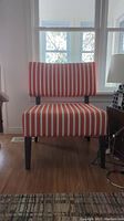 Front view of cushioned accent chair showing orange and beige vertical striped fabric and wood legs on laminate floor in front of windows.