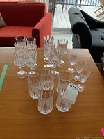 16 real crystal glasses arranged on table including multiple types: water glasses, large and small wine glasses, liqueur glasses. Visible cut patterns on glass surfaces.