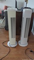 Two tower fans side by side on floor, showing white and yellowed plastic body and vertical vents.