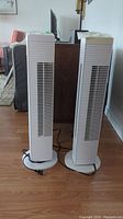 Two white Intertek tower fans side by side on a wooden floor with furniture in the background.