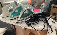 Photo showing three irons (two teal and white Proctor Silex, one white and green Philips) on tabletop ironing boards with hair styling tools.