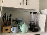 Overall view of kitchen shelf showing Cuisinart knife block, French press, two milk pitchers, pastel teapots, Proctor Silex blender and white serving tray.