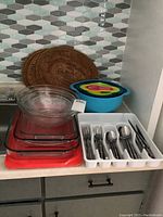 Full set of kitchenware items including glass casserole dishes and mixing bowls, plastic nested mixing bowls with measuring spoons, stainless steel cutlery set in white tray, and wicker placemats against backsplash.