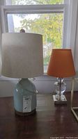 Two small table lamps side by side on wooden table. One lamp with light blue ceramic and wood base with off-white shade, the other lamp with clear glass base and orange shade.