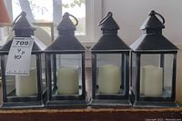 Photo showing all four battery operated candle lanterns lined up on a wooden surface, each with a black metal-look frame and cream battery candles inside.