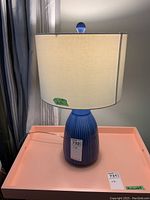 Blue ceramic table lamp with vertical ridged design and cream fabric shade, photographed on a pink surface against a neutral wall.