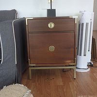 Front view of wooden nightstand with brass-tone metal accents and hardware, next to gray upholstered chair and white tower fan, showing scratches and wear.
