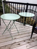 Two light green painted metal foldable patio side tables on a wooden deck near a railing overlooking water.