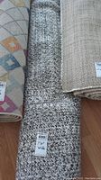 Rolls of rugs showing one braided grey area rug with label indicating 7'6" wide.