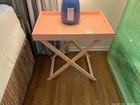 Pink folding tray table with blue lamp on top, wood floor, positioned near bed with striped bed skirt and curtains in background.