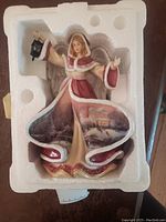 Figurine in original Styrofoam packaging showing angel in red and cream gown holding a lantern with winter village artwork on gown.
