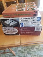 Three boxed items stacked on a table: indoor grill, double tier utensil drawer, Lazy Susan sectional server.