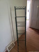 Foldable metal rack stand shown fully upright against wall, with decorative scrollwork at top and multiple wire grid shelves.