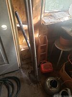 Photo showing pitch fork, shovel with long wooden handles, gas can near the ground, set inside a lighted shed corner.