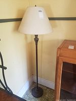 Full view of the tall floor lamp with metal stand and beige fabric lampshade on top, positioned near a beige wall and corner.