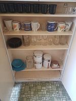 Shelves inside a white cupboard holding various mugs, glasses, small plates, jars, and a blue ceramic plate with attached cup.