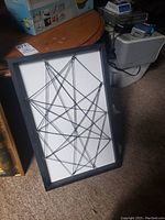 Black framed print featuring geometric design with intersecting black string on white background shown standing on carpet.