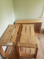 Two wooden slatted shelving units with natural finish, each with two slatted shelves, on worn wood floor against wall.