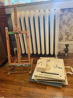 Wood easel, stack of papers/sketchbooks, part of a heating radiator in background, wooden floor