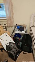 Photo shows white plastic bathtub seat leaning against a floral armchair, Dr. Ho's Perfect Backrests in black with massage nodes and red tips are placed on the floor, ObusForm backrest is visible to the right, and blood pressure monitor case with device on a small table in background.