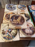 Photo showing seven collector plates arranged on original boxes. Plates depict nostalgic scenes of children in winter and indoor settings, with gold decorative rims.