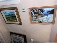 Photo showing two original framed paintings hanging on a wall, one coastal and one landscape.