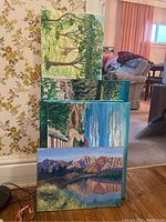 Five paintings stacked against a wall showing variety of green and blue landscape scenes.