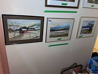 Three framed original paintings hanging on a wall showing various landscape scenes in snow, mountains and coastal views.