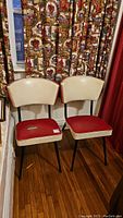 Pair of retro vinyl upholstered chairs with metal legs, shown side by side on wood floor with decorative curtain background.