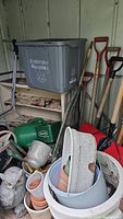 Shelf and floor view with grey plastic recycle bin, various gardening tools like seed spreader and watering can, plastic owl statue, planters stacked inside large plastic planters