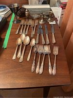 Full view of silver-plated cutlery including knives, forks, spoons, two candlestick holders, two wine goblets, salt shaker, coasters and napkin holder on wooden table.