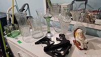 Full view of the lot showing multiple clear glass vases of various shapes and sizes, a green art glass vase with bubble detail, black metal frame holders, and a ceramic figurine of a woman reading newspaper.