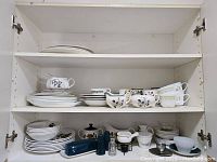 Photo showing multiple stacks of dinner plates, cups, and cream and sugar dishes on white shelving, displaying various floral and green bordered designs.