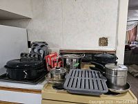 Overall assortment of pots, pans, baking and cooking ware arranged on kitchen counter including black enamel roasting pan, muffin tins, metal stock pot, steamer, double boiler, and cast iron grill pan.