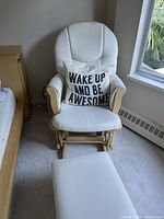 Front view of white maternity rocking chair with cushion and pillow that reads 'WAKE UP AND BE AWESOME'. Chair has wooden armrests and rocker base, placed near a window and bed.