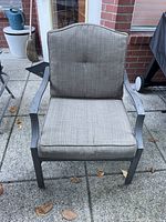 Outdoor metal framed chair with light brown seat and back cushions on a concrete patio with leaves scattered around.