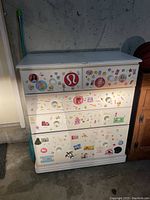 Full frontal photo of white pine dresser with four drawers covered in various colorful stickers including logos and cartoon images