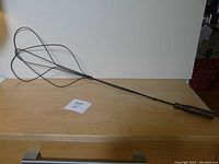 Full view of the antique carpet beater showing the long metal shaft and wooden handle laid on a wooden surface.