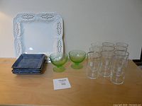 Full lot on table showing white lattice edged serving plate behind four blue pottery plates, two green glass dishes and ten clear Canadian Club glasses.
