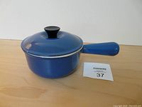Side view of the blue Le Creuset pot with lid and handle, displayed on wooden surface, showing intact lid and pot body with small chips on the rim.