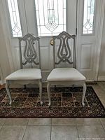 Pair of wooden side chairs positioned in front of a door showing full frontal view with beige upholstered seats and curved legs.