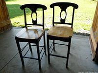 Front view of two wooden bar chairs showing blue painted frame and natural wood seat outdoors on a concrete floor.