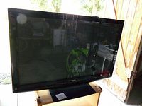Front view of Panasonic flat screen TV on stand, power cord draped over top, reflective screen showing background reflections.